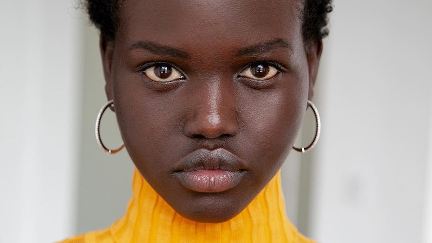 Adut Akech, wearing a yellow knitted top,  looks at the camera in a posed portrait.