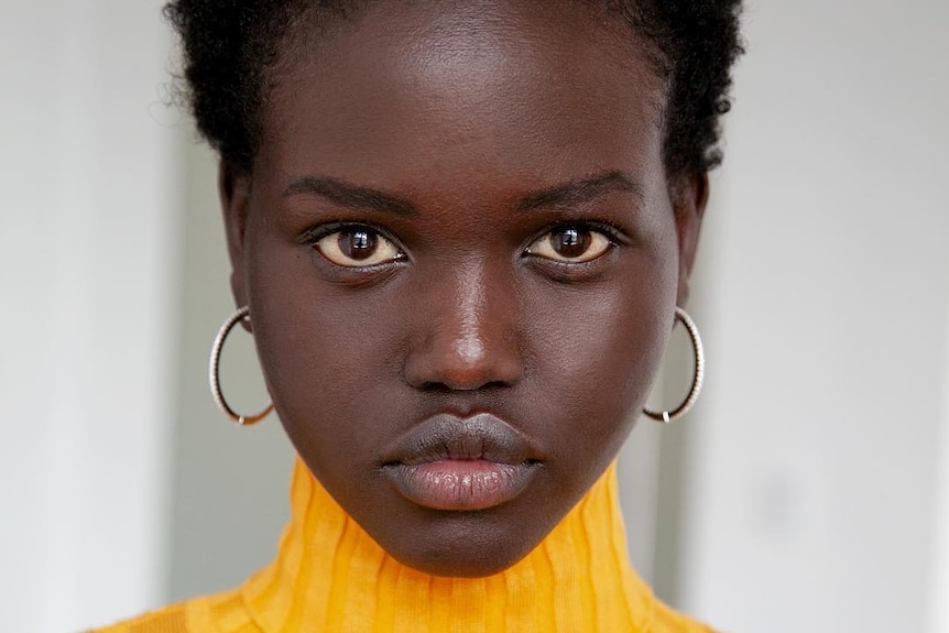 Adut Akech, wearing a yellow knitted top,  looks at the camera in a posed portrait.