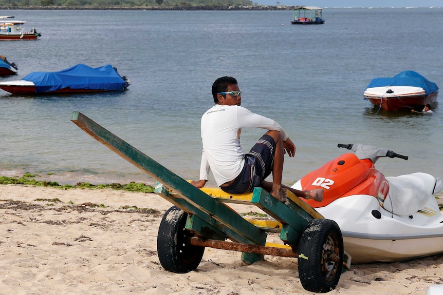 An Indonesian man with sunglasses sits on an empty beach with empty boats on the water.