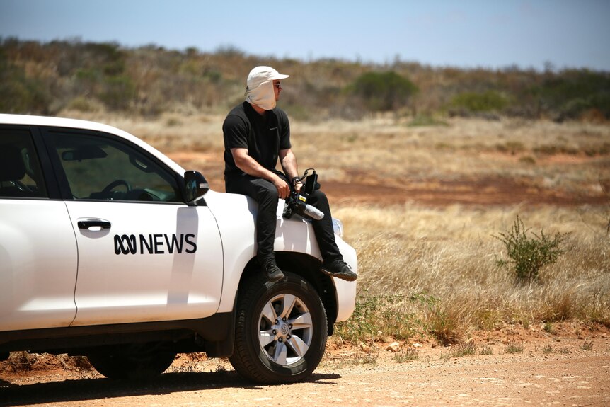 A man sits on the bonnet of an ABC News white four-wheel-drive holding a camera.