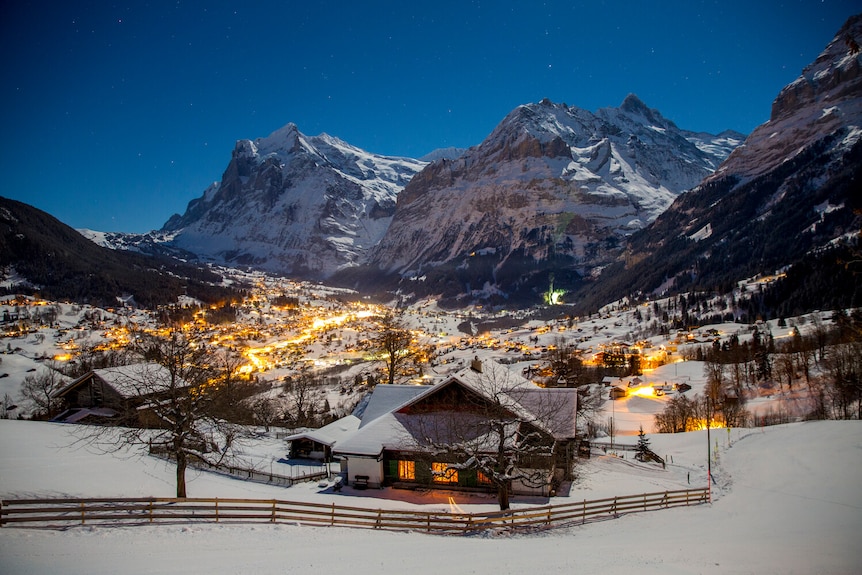 The Swiss village of Grindelwald at night cover in snow with towering mountains behind