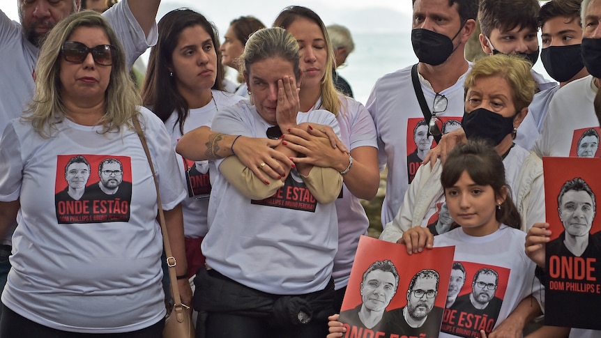 A crowd of people wear the same white shirt with the faces of the two men on them. Distraught women hug each other.