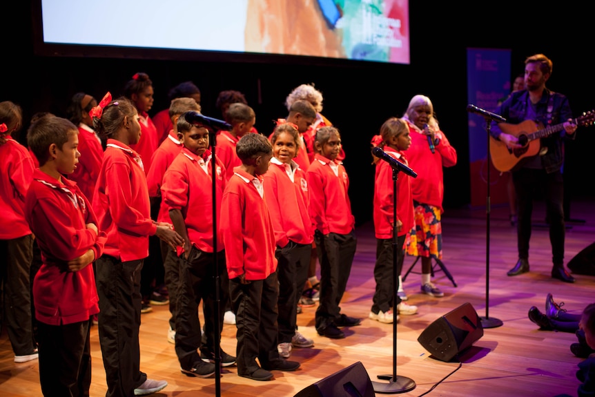 Students stand on stage at the Opera House singing along with a man on a guitar