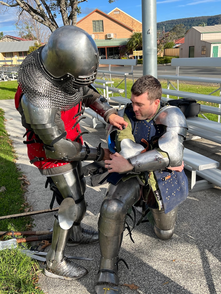 A man in knights armour & red coat helps a kneeling man in a blue coat put armour on his arm. 