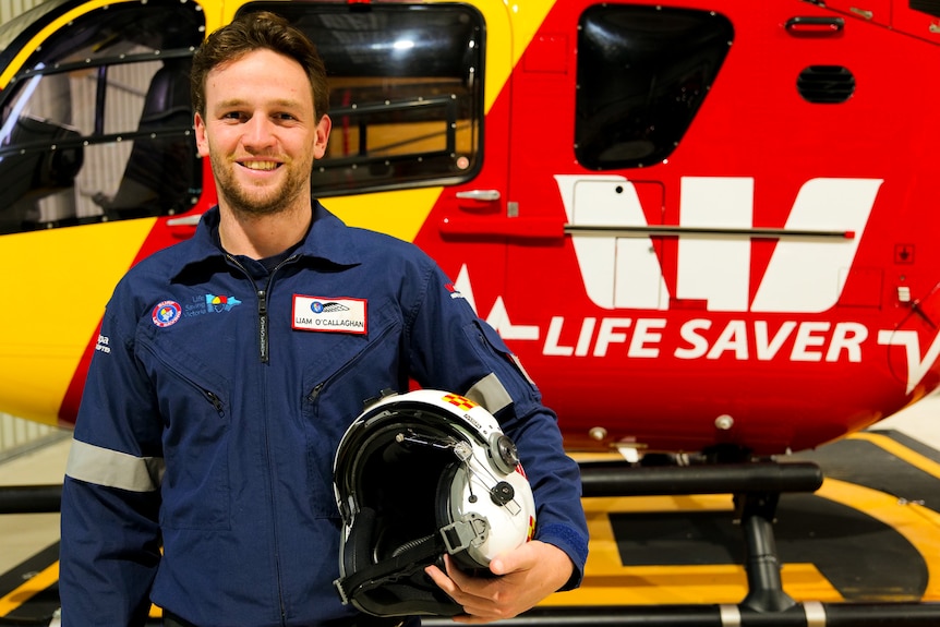 A man wearing navy blue overalls, and carrying a helmet, stands in front of a Westpac life saver helicopter. 