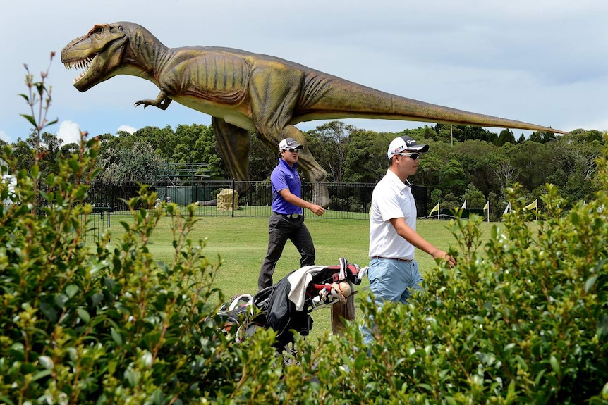 Golfers walk past a giant dinosaur statue on a course.