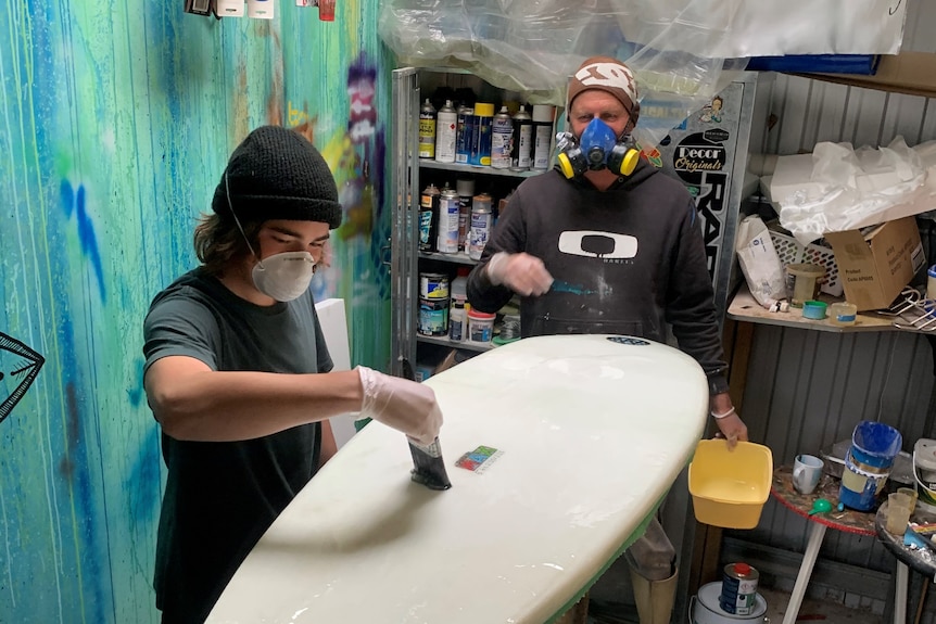 Man and boy wearing dust masks work on a fibreglass surfboard.