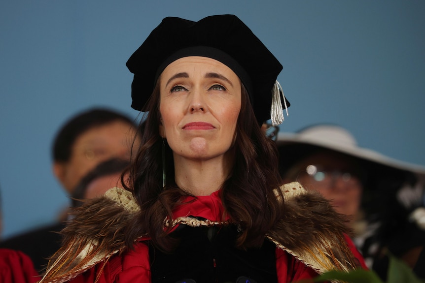 Jacinda Ardern wearing a black cap and red gown, looking up towards the sky