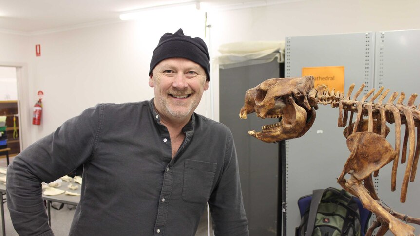 Julian Hume stands indoors next to a large animal skeleton, smiling.