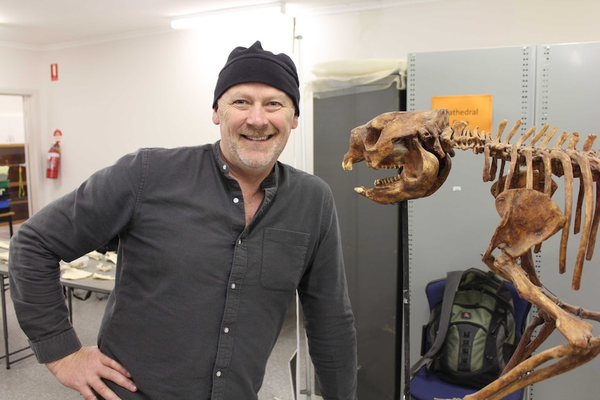 Julian Hume stands indoors next to a large animal skeleton, smiling.