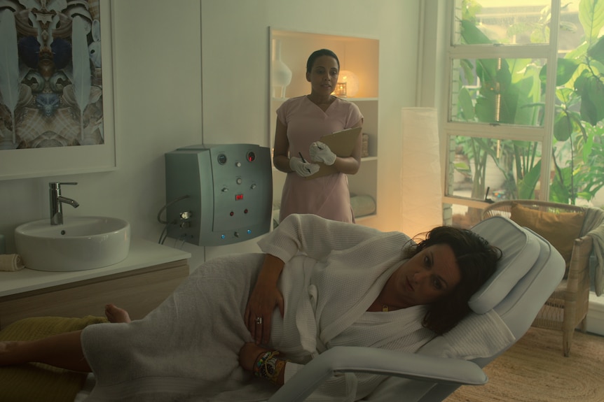 A 40 year old woman in a white dressing down lies down to the side having a colonic, a woman in white behind her, from a TV show