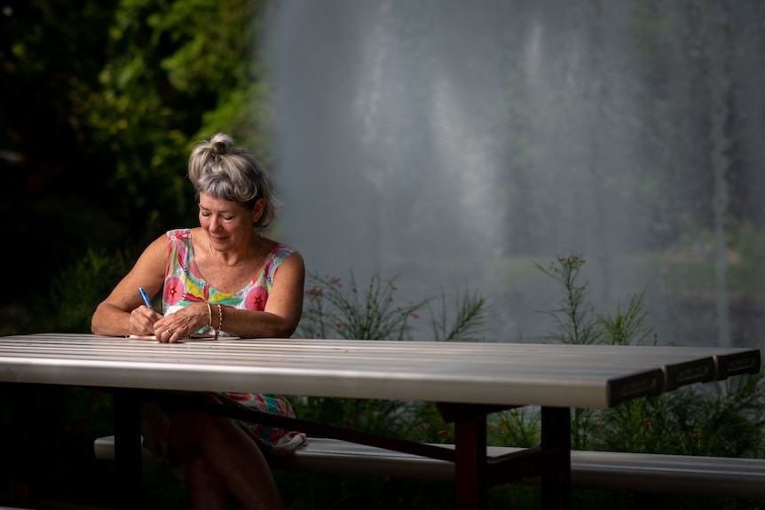 A woman sits on the end of a park bench and is writing. Behind her,  there's a fountain and a lush garden.