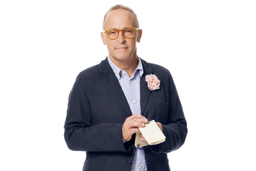 A photo of David Sedaris shows him holding a notebook, a flower on his lapel.