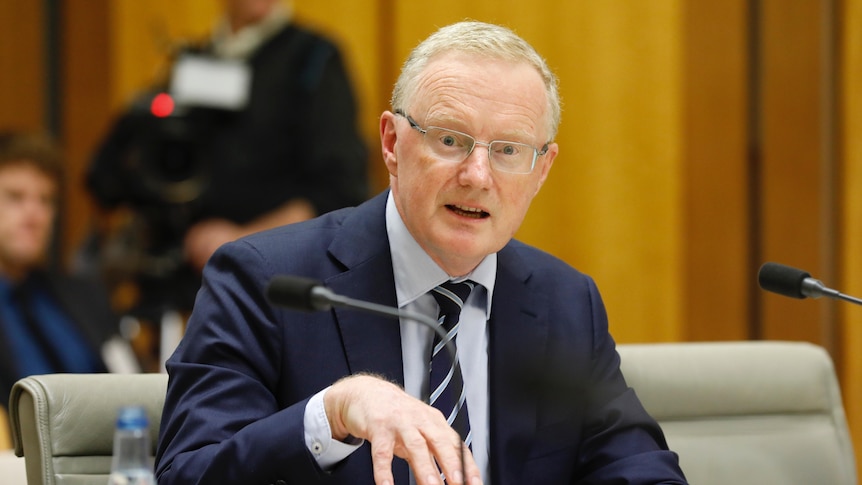 Philip Lowe sitting at a desk behind a microphone mid-sentence, gesturing with his right hand and looking at the camera