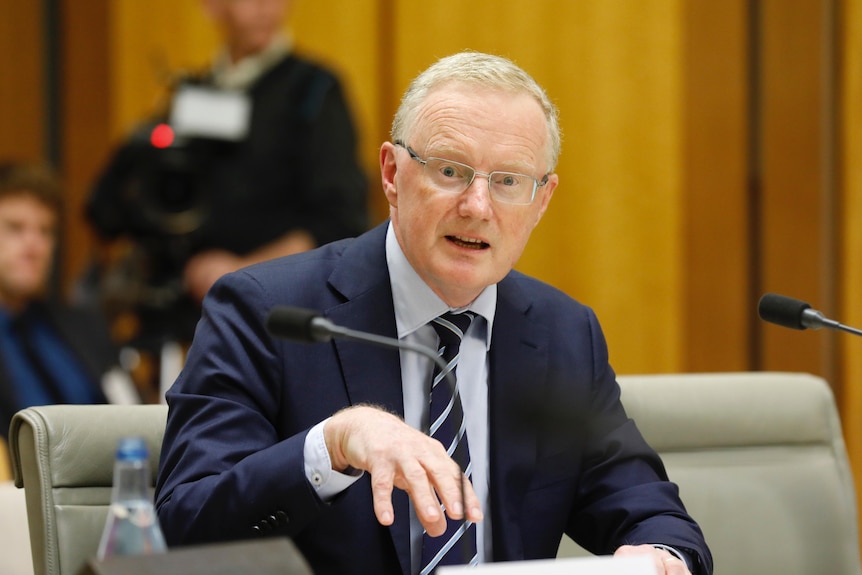 Philip Lowe sitting at a desk behind a microphone mid-sentence, gesturing with his right hand and looking at the camera