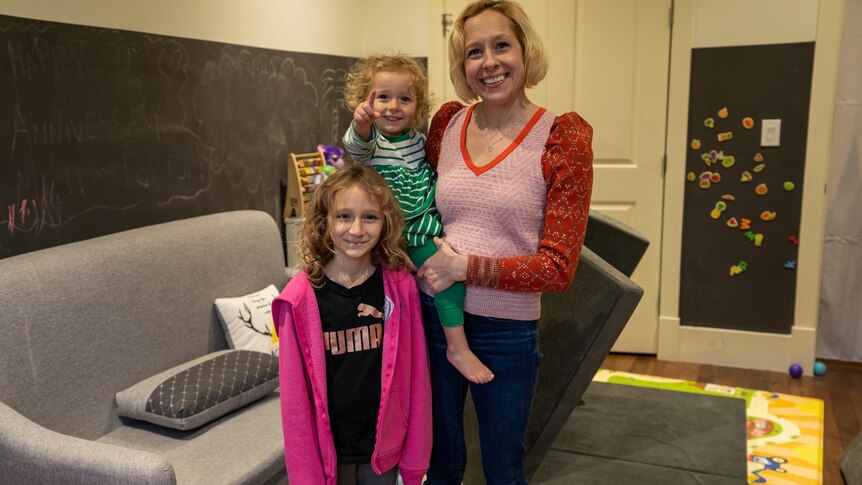 Woman stands with her two children in a living room. 