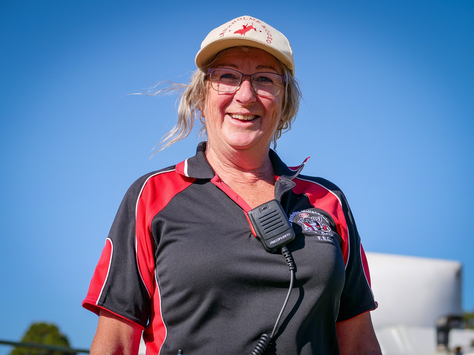 A woman wearing a rodeo branded cap, a football club polo and walkie talkie smiles at the camera. 
