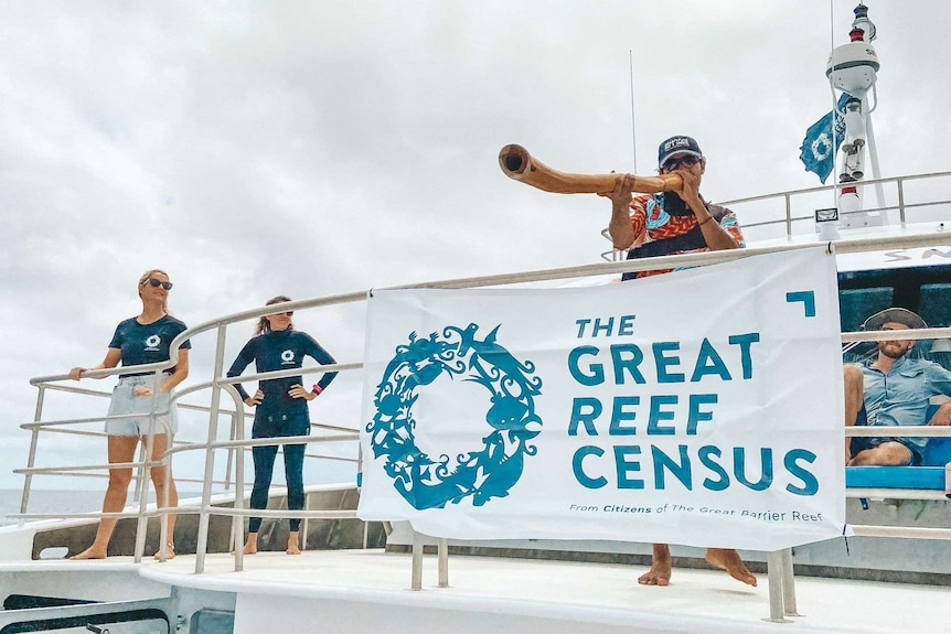 Man playing a didgeridoo on board a boat standing behind a "Great Reef Census" banner.