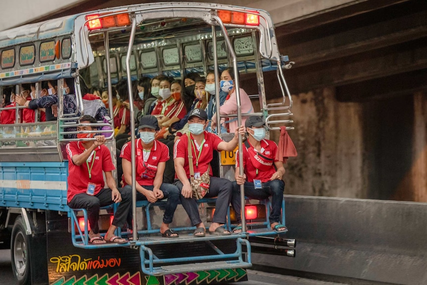 A group of people wearing masks sit on the back of a truck going down a street.