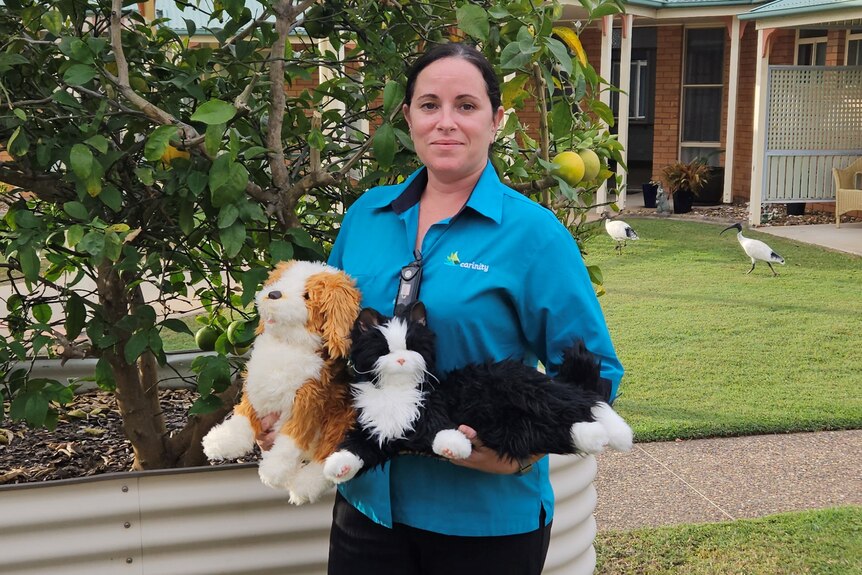 A woman in a blue button up stares at the camera holding two robotic pets, a garden behind her