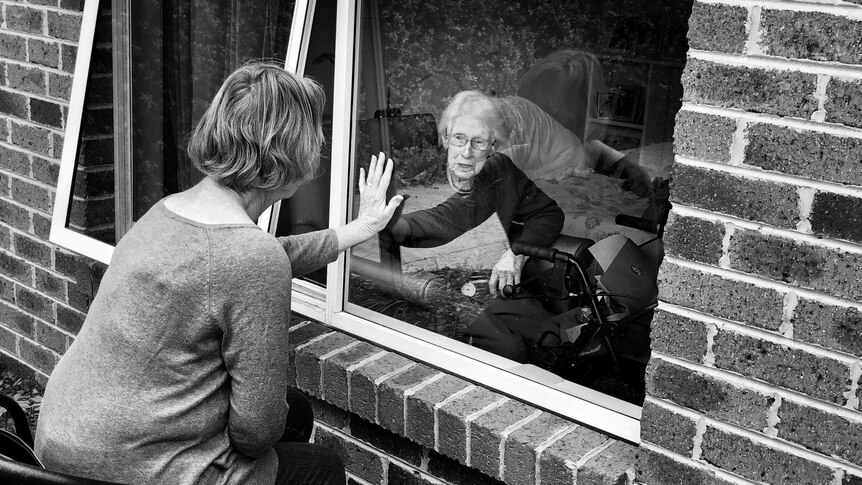A black and white image of two women, one elderly, placing their palms together on the window that separates them.