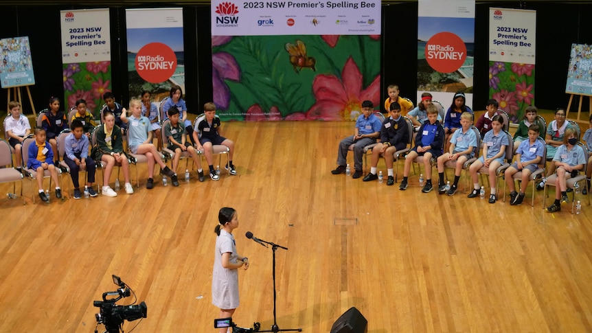A girl stands at a microphone in front of a crowd of seated children