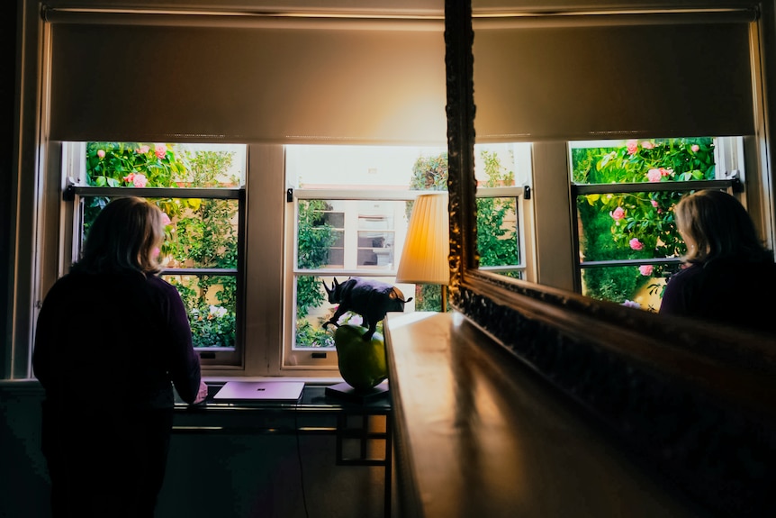 a woman standing by the window in a dark room, scene reflected by a wall mirror