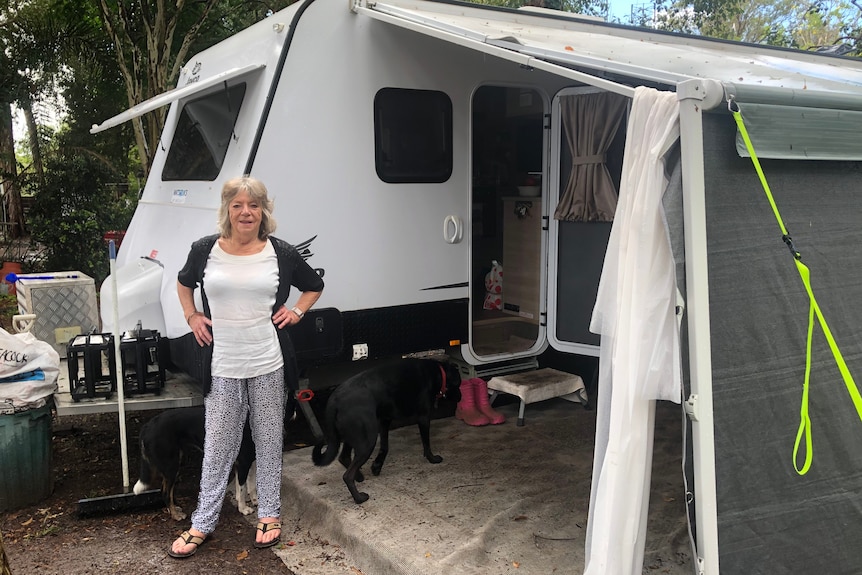 A middle-aged woman stands with hands on her hips outside a caravan with an awning. 