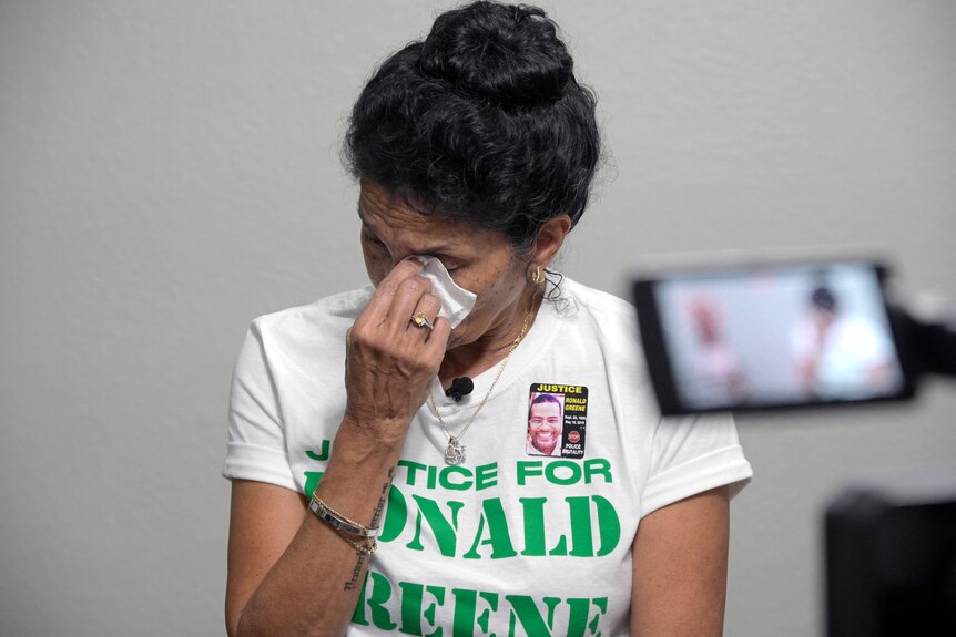 A woman with her hair in a bun is pictured crying while wearing a t-shirt that says 'Justice for Ronald Greene' 