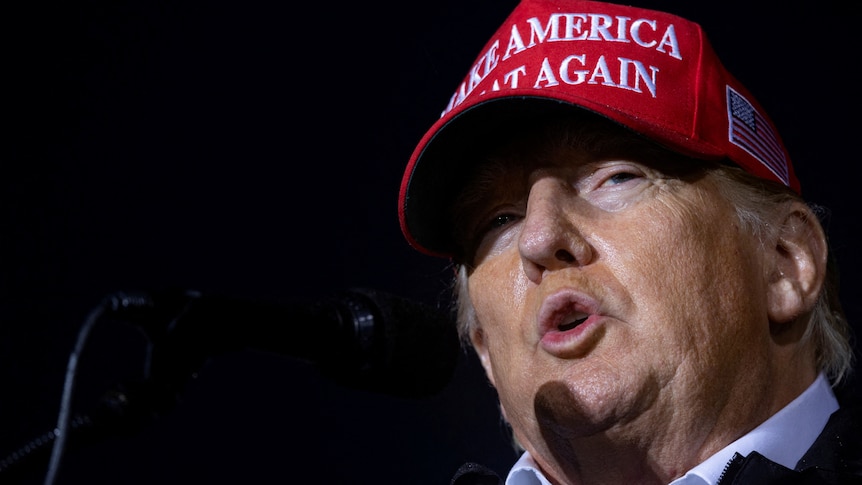 Donald Trump wearing a red 'make America great again' cap while pursing his lips 