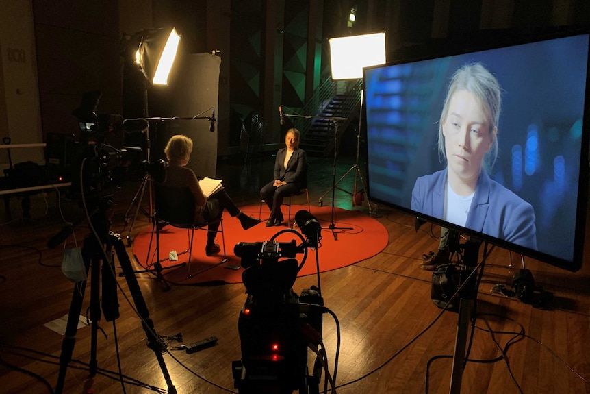 Wide shot of two women sitting on chairs surrounded by camera and lighting equipment and large TV screen.