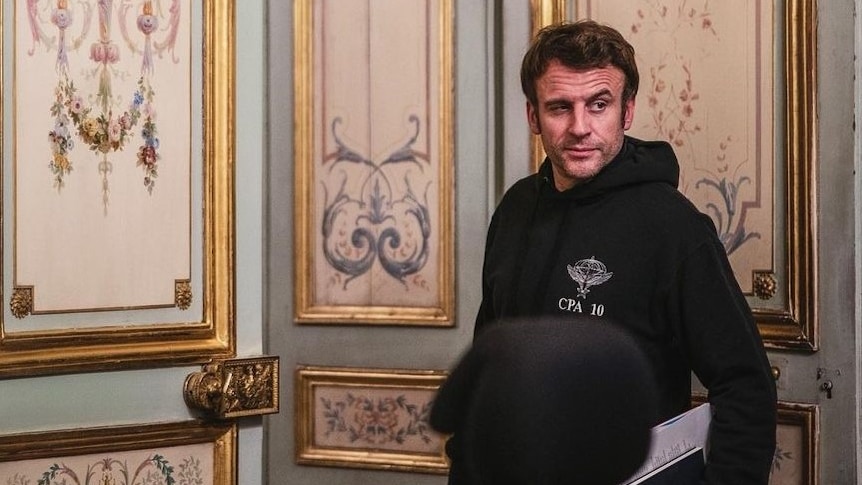 Emmanuel Macron stands in front of a decorated wall wearing a hoodie.