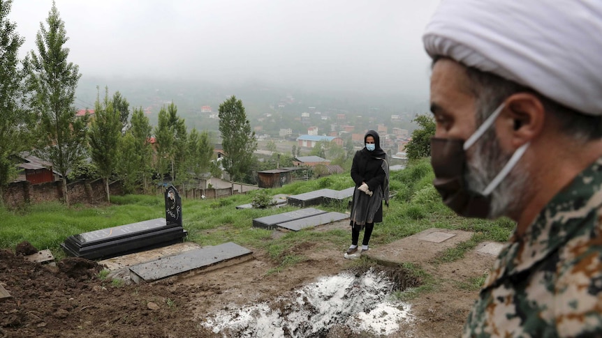 A man wears a mask while standing in a graveyard as a woman wearing a mask and gloves, prays at the grave behind him.
