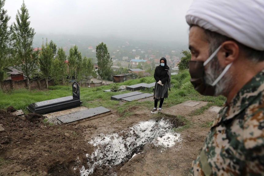 A man wears a mask while standing in a graveyard as a woman wearing a mask and gloves, prays at the grave behind him.