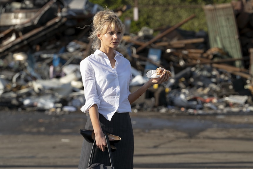 Young woman with long light blonde hair in 9-5 work clothes holding heels and bag in one hand and hot dog in other, outside.