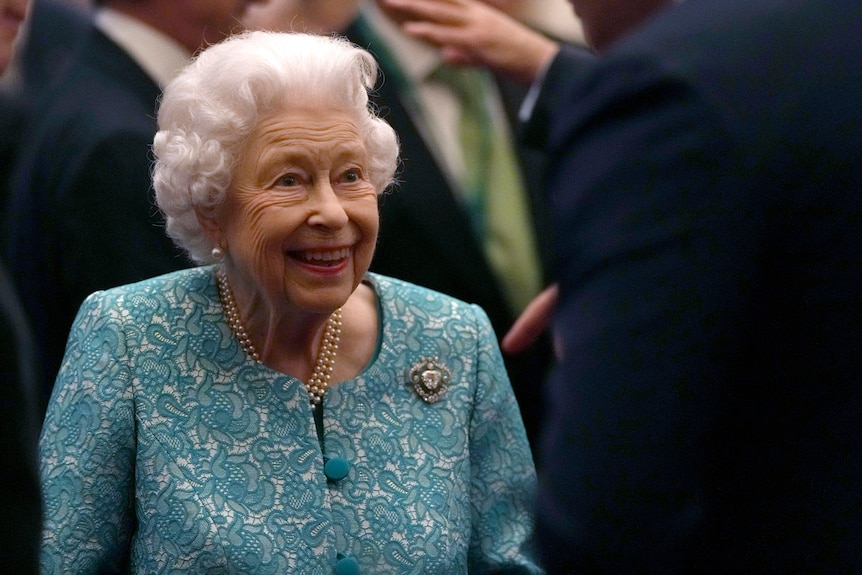 The Queen in a blue lace blazer and pearls, smiling