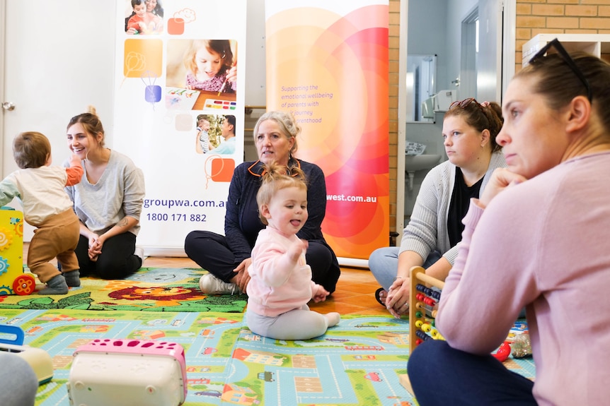 A group of women sit on the floor in a circle with some small children with them.