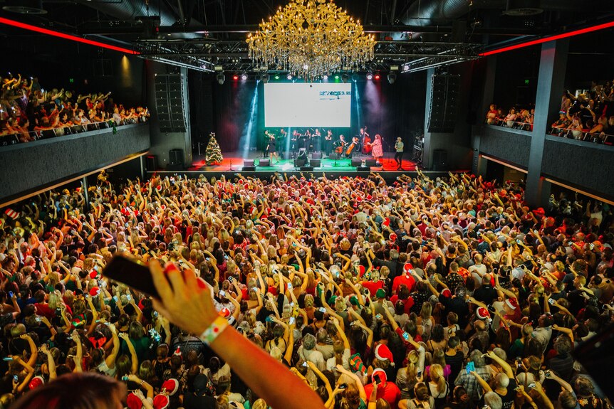 A theatre interior with hundreds of people waving their mobile phones as musicians perform on a stage.