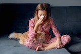 Girl in pajamas on coach, blowing her nose with a long-haired cat on her lap