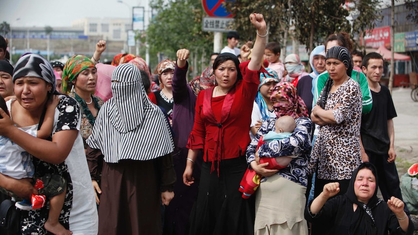 Women in scarves stand, woman in centre wears red and puts her fist in the air. Another woman holds a baby.