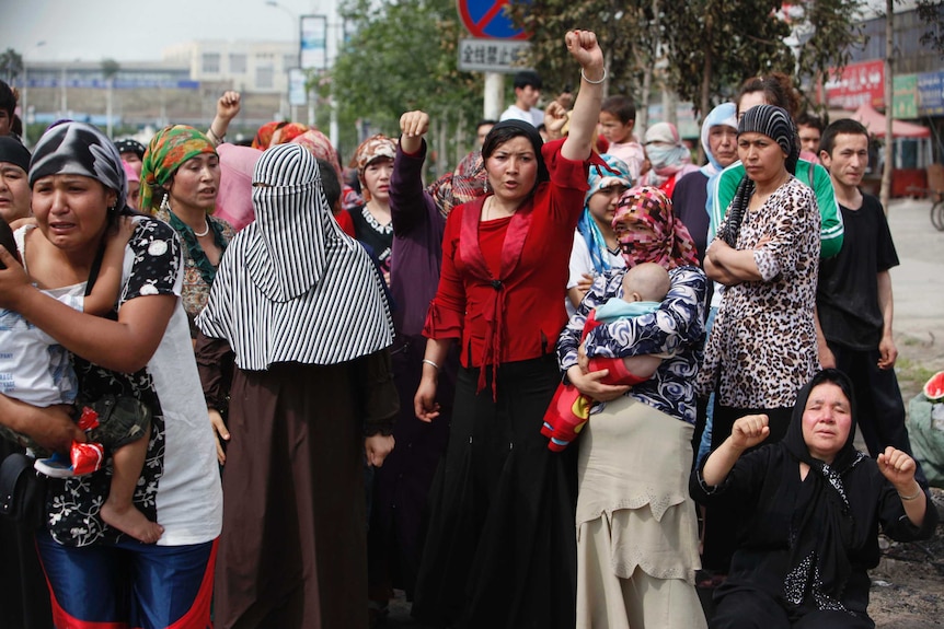 Women in scarves stand, woman in centre wears red and puts her fist in the air. Another woman holds a baby.