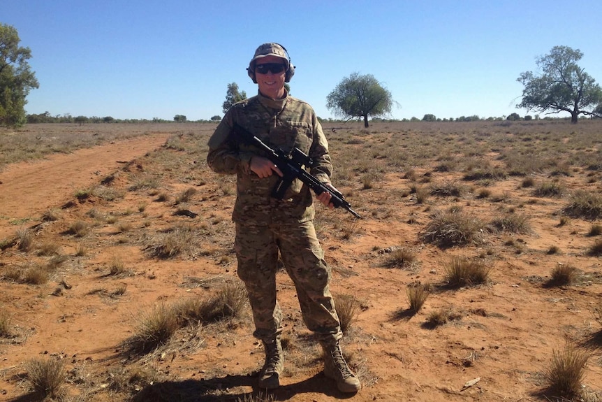 Dressed in camouflage, Ozzie Dixen holds a rifle while standing to the side of a dirt road on an outback Queensland property.