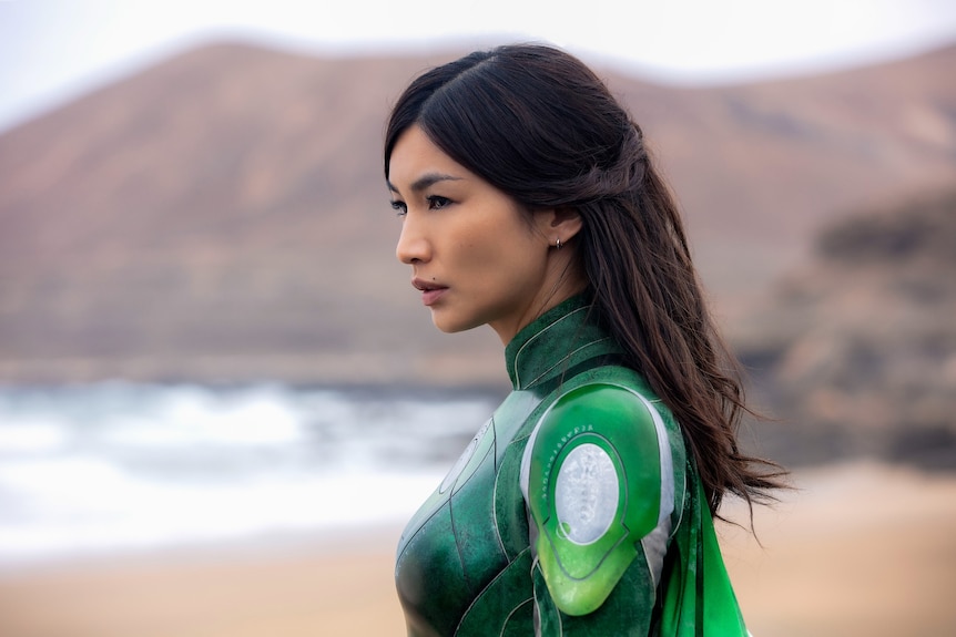 Gemma Chan, wearing a green superhero costume, looks determined as she stares into the distance on a beach