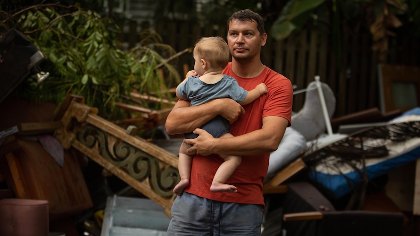 A man stands holding a baby in front of a pile of ruined household items.