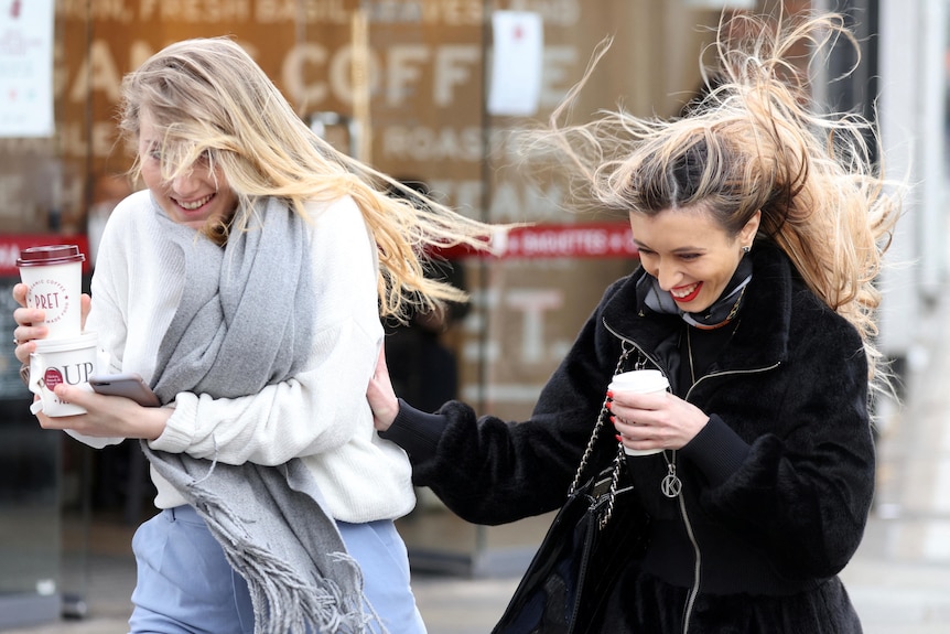 Two women hold coffee cups, their hair blowing in the wind.