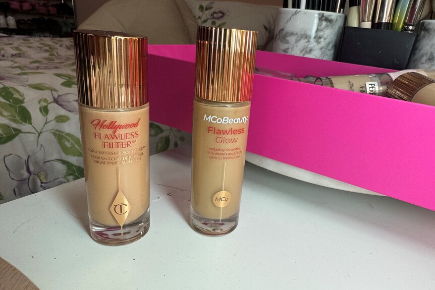 Two nearly identical cylindrical bottles with gold lids sit on a while desk.
