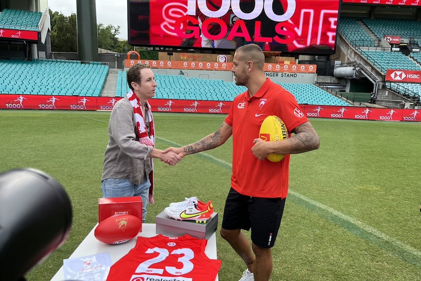 Sydney Swan Lance Franklin shakes the hand of a Sydney fan while holding a football - in the background is a sign "1000 goals".