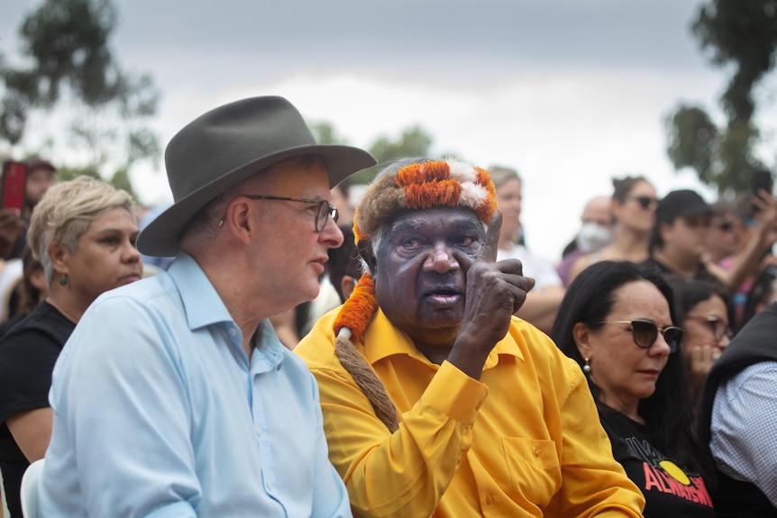 Two men sitting next to each other and talking, with one holding up a hand as he speaks. 