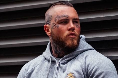 A muscular man with tattoos on his face leans against a wall.