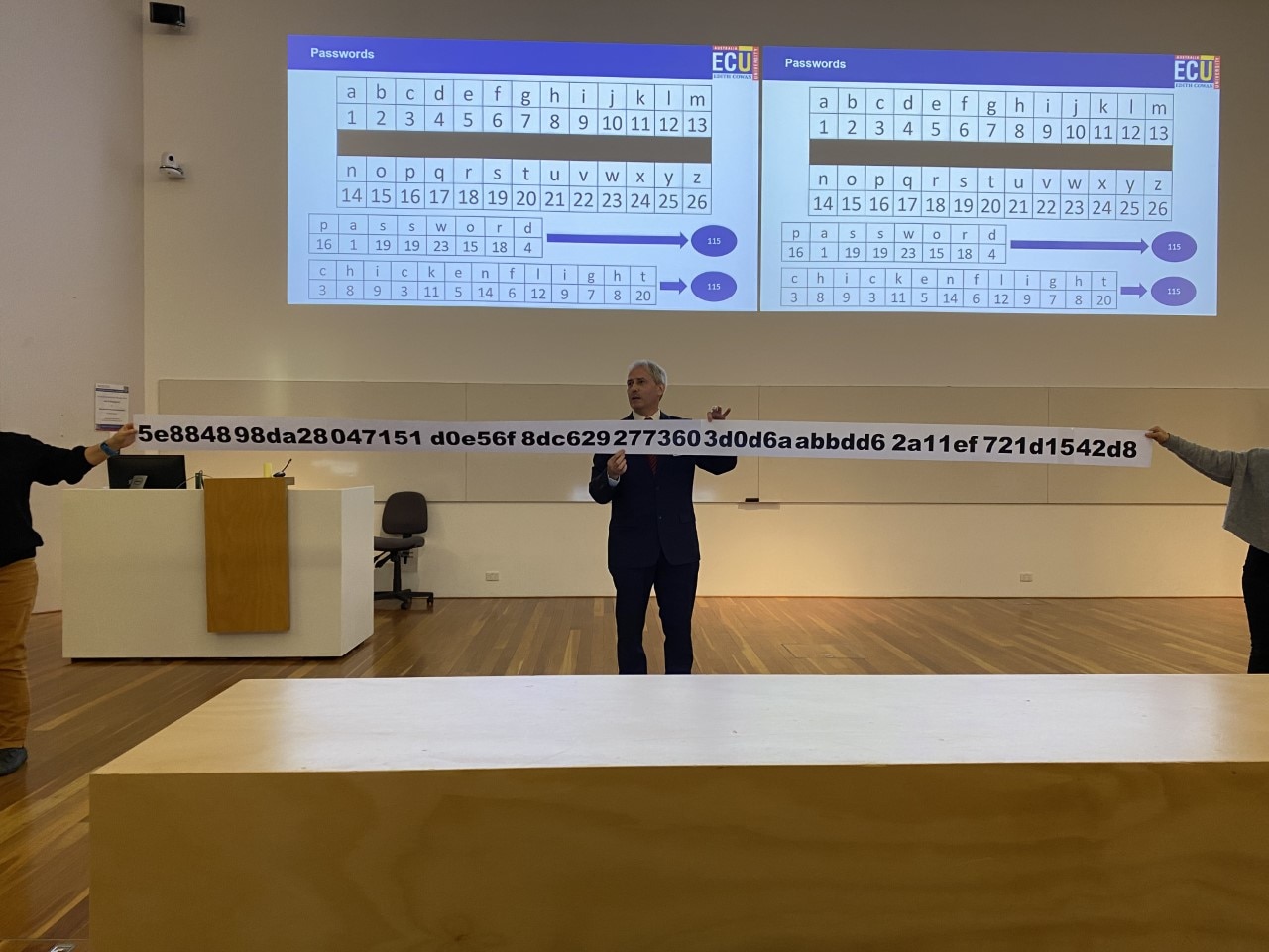 A man in a suit giving a lecture holds a ridiculously long piece of paper containing a password of random letters and numbers.